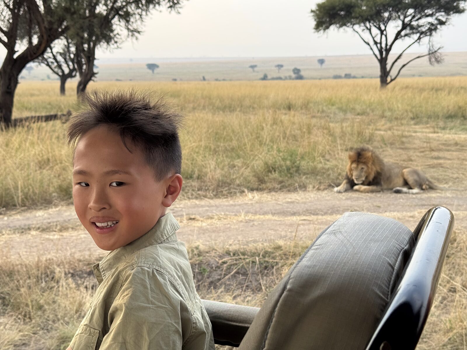 Our son grinning from the safari vehicle with a male lion lying beside the track behind him