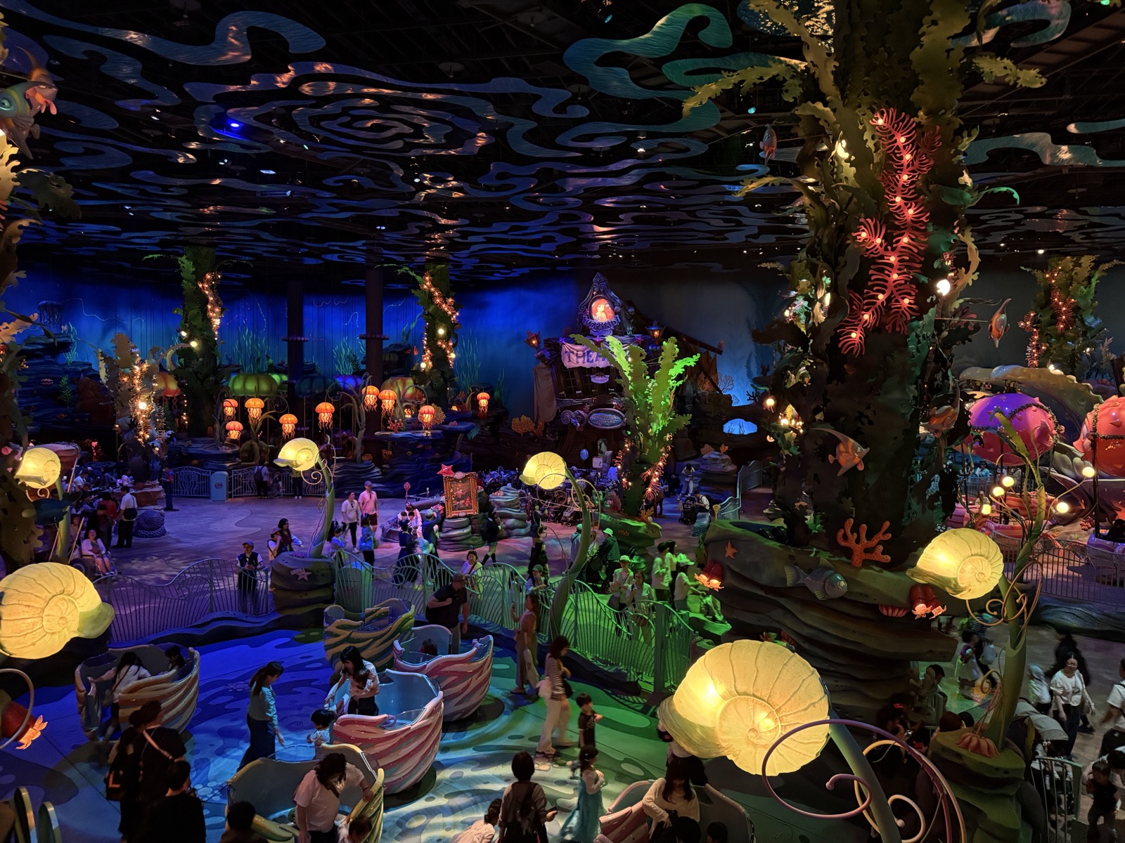 Inside Mermaid Lagoon at Tokyo Disney Sea — a covered indoor plaza lit by glowing jellyfish lanterns, with a coral-and-seaweed canopy and small carnival rides below
